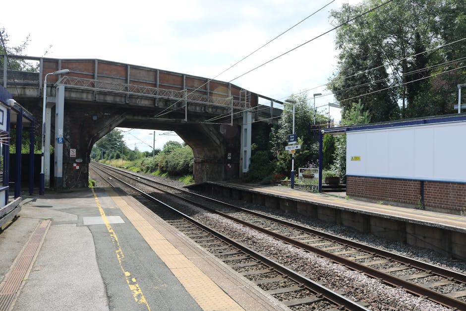 A quiet railway platform adjacent to the tracks, featuring a yellow safety line along the edge, small metal benches, and a modern lamppost. Overhead, a concrete bridge with geometric cut-outs spans ac